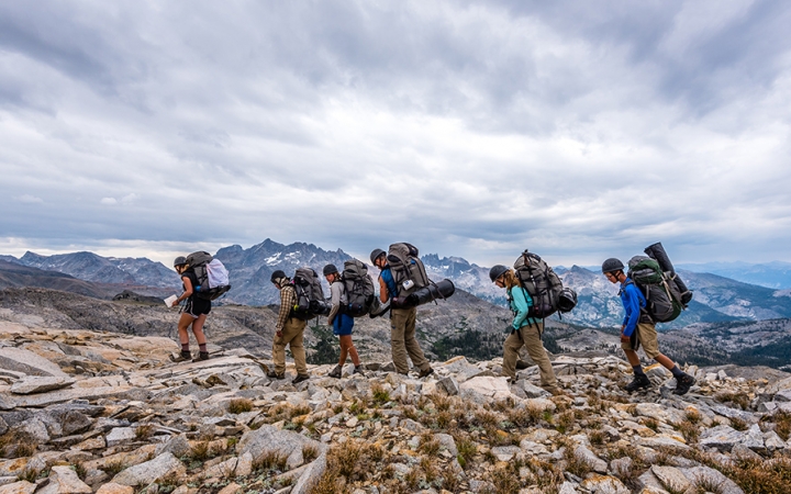 A group of students wearing backpacks high across rocky terrain with mountains in the background. 